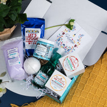 Load image into Gallery viewer, Assorted self-care products including bath bombs, soap, and body wash in a box on a yellow textured surface.