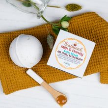 Load image into Gallery viewer, Oatmeal Milk &amp; Honey bath bomb with packaging on a mustard-colored towel.