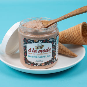 Jar of 'a la mode' whipped soap scrub with a wooden spoon on a blue background