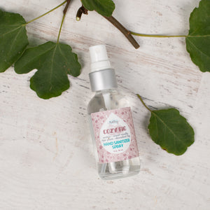 Hand sanitizer spray bottle with a pink label on a light wooden surface with green leaves.
