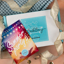 Load image into Gallery viewer, A Bath Bevy Relaxation gift box set styled on a tan surface with a blue pom-pom bath mat and gold ribbon. The set includes a white box with a turquoise logo and a colorful 'Summer party' invitation card featuring a sunset gradient and palm trees.