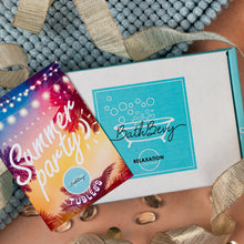 Load image into Gallery viewer, A Bath Bevy Relaxation 'Summer Party!' subscription box set styled on a peach-colored surface with a blue pom-pom bath mat and gold ribbon. The set includes a colorful tropical-themed card and the white box with a teal logo.