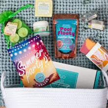 Load image into Gallery viewer, A curated summer spa and party gift basket arranged in a white woven basket on a gray textured blanket. The bundle includes a colorful 'Summer Party' pouch, a 'Pool Floaty' seaweed pack, a jar of 'Ankahuhi' body butter, a Lemon Grass steamer cube, tea lights in a green bag, and other small spa items.