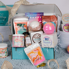 Load image into Gallery viewer, A curated bath and body gift box filled with assorted products including bath bombs, bath salts, whipped soap, arranged in and around a turquoise gift box with a beige ribbon on a blue textured bath mat.