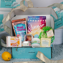 Load image into Gallery viewer, An open teal gift box filled with summer-themed bath and body products, including a colorful 'Pool Floaty' pouch, a 'Summer party' card, a green coconut lemon soap bag, a lemon, and other packaged items, set on a blue knit blanket with teal branded packaging in the background.