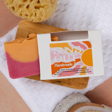 Load image into Gallery viewer, A handmade soap bar with a pink and orange gradient, displayed next to its white packaging box labeled 'SUNS OUT Handmade Soap' on a wooden board over a white towel.