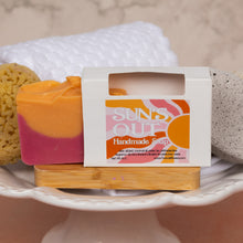 Load image into Gallery viewer, A handmade soap bar with a two-tone orange and pink color, displayed next to its white cardboard packaging box labeled 'SUNS OUT Handmade Soap'. The soap and box are presented on a wooden block atop a white ceramic pedestal, surrounded by natural bath accessories like a yellow sponge and a gray stone.