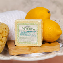 Load image into Gallery viewer, A square package of Lemongrass Shower Steamer Cube sits on a small wooden board, surrounded by lemons and a sponge on a white dishcloth background.