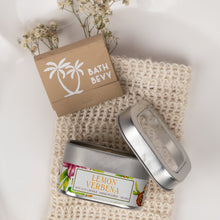 Load image into Gallery viewer, A silver metal tin of Bath Bevy Lemon Verbena bath salts sits on a cream-colored knit towel, with its kraft paper box and dried baby's breath flowers nearby, all arranged in a clean, flat-lay composition.