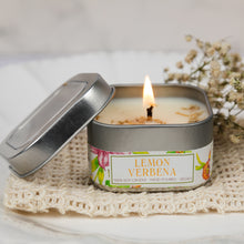 Load image into Gallery viewer, A lit soy candle in a square metal tin with the lid leaning against it, placed on a cream crocheted cloth with dried flowers in the background. The label reads 'LEMON VERBENA' and '100% SOY CANDLE - HAND POURED - VEGAN'.