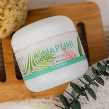 Load image into Gallery viewer, A white jar of Awapuhi Body Butter with tropical green and pink label graphics, styled on a wooden soap dish with a sponge, eucalyptus leaves, and a textured towel.