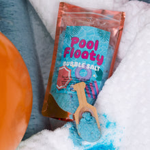 Load image into Gallery viewer, A pouch of Pool Floaty Bubble Salt with a small wooden scoop of the bright blue salt, styled on a white textured towel with a beach ball partially visible.