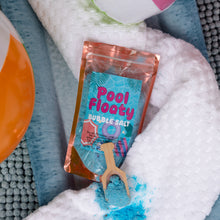 Load image into Gallery viewer, A pouch of Pool Floaty Bubble Salt is shown on white textured towels with blue pool water and a wooden scoop of the blue salt below it.