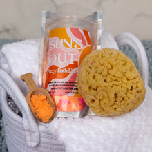 Load image into Gallery viewer, A pouch of SUN'S OUT Fizzy Bath Dust bath bomb powder sits in a white woven basket with a yellow sponge and a small wooden scoop filled with orange bath powder.