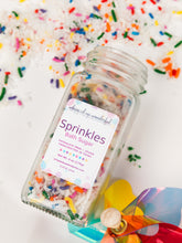 Load image into Gallery viewer, A clear glass jar of Whipped &amp; Wonderful 'Sprinkles Bath Sugar' filled with white sugar and multicolored sprinkles, shown on a white background with scattered sprinkles and a colorful heart-shaped prop.