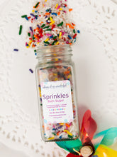 Load image into Gallery viewer, A clear glass of Whipped Up Wonderful Sprinkles Bath Sugar filled with multicolored bath sugar sprinkles, shown on a white lace doily with more sprinkles spilled above and colorful ribbons at the bottom.