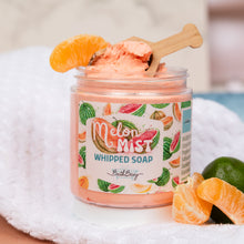 Load image into Gallery viewer, A jar of Bath Bevy Melon Mist Whipped Soap sits on a white towel, surrounded by melon and lime slices. The clear jar shows the pink whipped soap inside, with a wooden spoon resting in it.