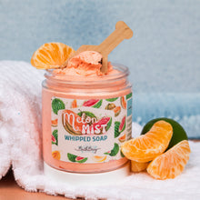 Load image into Gallery viewer, An open jar of Melon Mist Whipped Soap by Bath Bevy, filled with peach-colored whipped soap and topped with a wooden spoon, set on a white towel with orange slices and a lime against a light blue background.