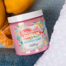 Load image into Gallery viewer, A jar of Bath Bevy Watermelon Lemonade Whipped Soap sits on a textured blue surface next to a white towel, with lemons and orange slices as props.
