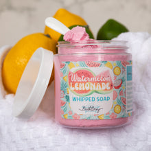 Load image into Gallery viewer, A jar of Bath Bevy Watermelon Lemonade Whipped Soap sits on a white towel, with lemons and a lime behind it as props. The open jar shows pink whipped soap inside, with the lid leaning against the left side.