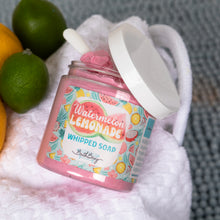 Load image into Gallery viewer, A jar of Bath Bevy whipped soap labeled Watermelon Lemonade, shown open with pink whipped soap inside and a white lid leaning against it on a white towel. Limes and citrus slices are placed around it, with a gray textured background.