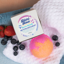 Load image into Gallery viewer, A colorful bath bomb with pink and orange layers sits on a white towel next to its white packaging box labeled 'Bikini Blast' and surrounded by strawberries and blueberries.