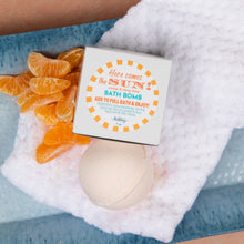 Load image into Gallery viewer, A bath bomb package labeled 'Here comes the SUN Bath Bomb' placed on a white textured towel with orange mandarins and a pale bath bomb visible underneath on a blue surface.