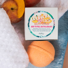 Load image into Gallery viewer, A peach-colored bath bomb sits on a blue textured surface next to a white towel, with sliced peaches and the product box. The box label reads 'TROPICAL PEACH BATH BOMB' with ingredients listed below.