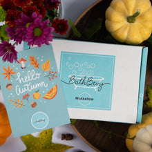 Load image into Gallery viewer, A BathBevy 'Relaxation' bath box displayed next to an autumn-themed 'hello autumn' insert card. The scene is styled with fall flowers, acorns, and small pumpkins on a dark wooden tray.