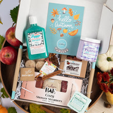 Load image into Gallery viewer, An autumn-themed gift box filled with bath and beauty items, including a Hygge body wash, a 'hello autumn' card, a lavender pajamas scented candle, Cozy Shower Steamers, and a Bath Bevy soap bar, arranged in a wooden bowl with apples, leaves, and a small pumpkin.