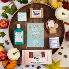 Load image into Gallery viewer, A styled flat-lay of multiple bath and beauty products arranged on a round wooden tray, surrounded by fall-themed props including pumpkins, leaves, apples, and cinnamon sticks. Visible products include a teal bottle labeled Happy Berry Body Wash, a lavender pajamas whipped soap scrub, a pink box of RAE Cozy Shower Steamers, a blue card reading hello autumn, and other packaged items.