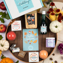 Load image into Gallery viewer, A styled autumn gift set from Bath Bevy, featuring a wooden tray with a 'Hello Autumn' card, a bottle of Bubble bath, a bag of Harvest Blend bubble salt, and various scented items, surrounded by apples, pumpkins, and flowers on a light wood background.