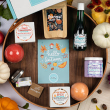 Load image into Gallery viewer, A styled flat-lay of a fall-themed gift basket or product assortment arranged on a round wooden tray. Visible items include a blue 'hello, autumn' card, a bag labeled 'Harvest Bubble salt', a bottle labeled 'Hygge happy', a candle labeled 'crisp Apple', a bath bomb box labeled 'Pumpkin Spice', a jar labeled 'Lavender pajamas', and other small seasonal products, with pumpkins, leaves, and dried flowers around them.