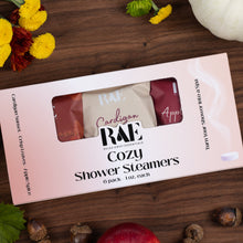 Load image into Gallery viewer, A boxed pack of RAE Health &amp; Wellness Cozy Shower Steamers sits on a dark wood surface, surrounded by autumn-themed props including yellow flowers, green leaves, acorns, and a white pumpkin. The pale pink box shows three visible packet colors: rose, cream, and burgundy, with the brand name RAE and product text clearly visible.