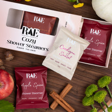 Load image into Gallery viewer, RAE Cozy Shower Steamers gift set displayed on a rustic wooden surface with autumn props including an apple, cinnamon sticks, acorns, and a small pumpkin. The set includes a pink outer box with a clear window, a cream resealable packet labeled Cardamom Sunset, and two dark red packets labeled Apple Spice.