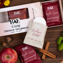 Load image into Gallery viewer, RAE Relaxation Essentials shower steamers arranged on a wooden surface with an apple, cinnamon sticks, and greenery. Visible products include a boxed Cozy Shower Steamers pack, a single Cardigan Sunset packet, and two Apple Spice packets.