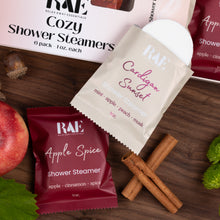 Load image into Gallery viewer, Three RAE Relax Away Essentials shower steamers arranged on a dark wood surface: a white box labeled Cozy Shower Steamers in the back, a cream packet labeled Cardiganum Sunset in the center, and a deep burgundy packet labeled Apple Spice in the foreground. A peach, cinnamon sticks, mint leaves, and acorns are placed around them as props.