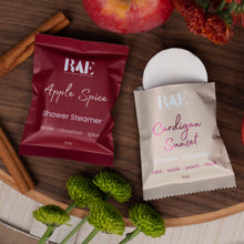 Load image into Gallery viewer, Two RAE Shower Steamer packets, one Apple Spice in a dark red packet and one Cardigan Sunset in a beige packet, styled on a wooden tray with apples, cinnamon sticks, and green flowers.