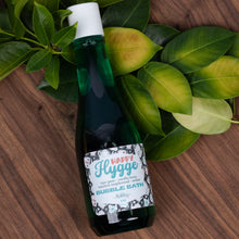 Load image into Gallery viewer, A dark green bottle of 'Happy Hygge Bubble Bath' with a white cap, styled on a wooden surface with green leaves.