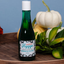 Load image into Gallery viewer, A dark green glass bottle of Happy Hygge Bubble Bath with a white cap, set on a wooden tray with white pumpkins and green leaves against a blue background.