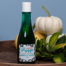 Load image into Gallery viewer, A green glass bottle of Happy Hygge Bubble Bath with a white cap, set on a wooden tray with white pumpkins and green leaves against a blue background.