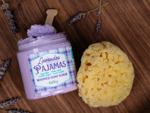 Load image into Gallery viewer, Lavender Pajamas whipped soap scrub in a clear jar with a wooden spoon, shown next to a yellow sponge on a wooden surface with lavender sprigs.