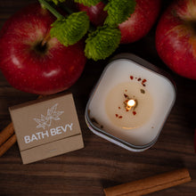 Load image into Gallery viewer, A lit candle in a square glass container sits on a dark wooden surface surrounded by red apples, green leaves, cinnamon sticks, and a branded matchbox-style box.