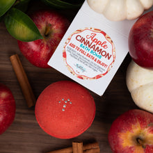 Load image into Gallery viewer, An Apple Cinnamon Bath Bomb set displayed on a dark wooden surface, featuring a red bath bomb, its white packaging box with a colorful label, whole red apples, cinnamon sticks, and small white pumpkins.