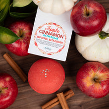 Load image into Gallery viewer, A square white bath bomb box labeled 'Apple Cinnamon Bath Bomb' is arranged on a wooden surface with whole red apples, cinnamon sticks, green leaves, and small white pumpkins around a round red bath bomb.