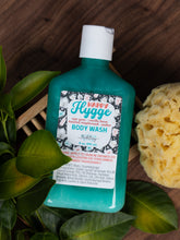 Load image into Gallery viewer, A teal bottle of Happy Huggge Body Wash with a white cap, styled on a dark wood background with green leaves and a yellow sponge.