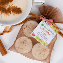 Load image into Gallery viewer, Four round soy candles in a sheer brown gift bag with a pink ribbon and a 'Green Daffodil Cinnamon Chai' tag, styled next to a mug of hot chai with cinnamon on a white background.