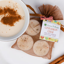 Load image into Gallery viewer, A styled product shot of Green Daffodil Cinnamon Chai hand-poured soy candles. The image shows a white bowl filled with a creamy beige candle topped with cinnamon powder, alongside several round candles in a sheer brown bag tied with a pink ribbon and a product tag, with cinnamon sticks on a white background.