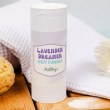 Load image into Gallery viewer, Body powder container labeled 'Lavender Dreamer' on a wooden surface with a towel and flowers in the background.