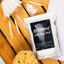 Load image into Gallery viewer, Package of 'Unwind Bath Fizzy Dust' with a yellow bath sponge on a wooden surface.